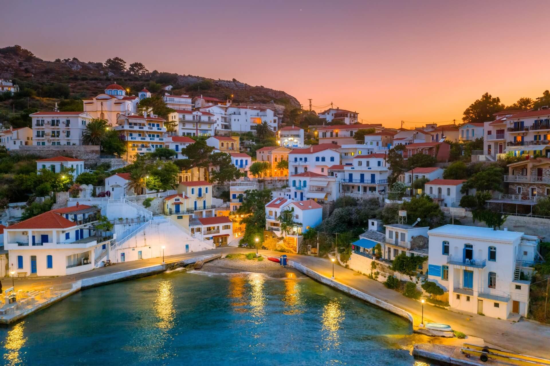 A tranquil Aegean village on Ikaria island at sunset, with whitewashed buildings against a backdrop of rolling hills and calm sea waters.