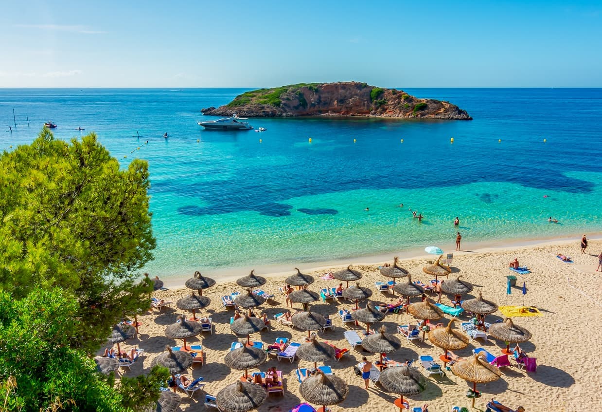 Strand von Mallorca, Spanien, mit türkisblauem Wasser, Sonnenschirmen aus Stroh, Badegästen und einer kleinen Insel im Hintergrund.