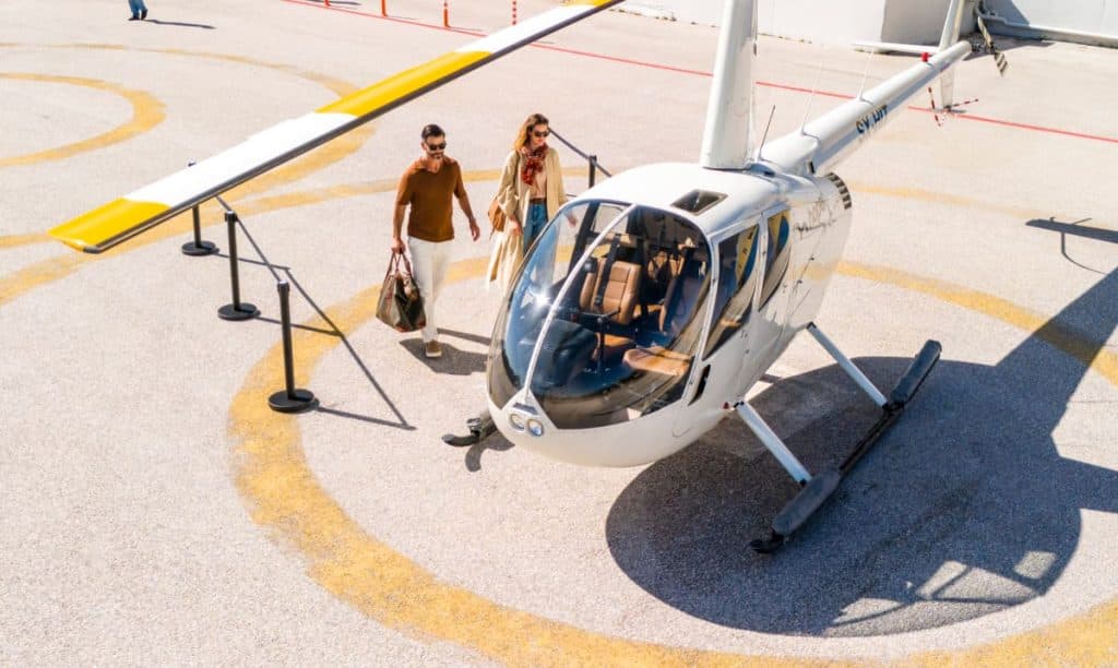 A stylish couple walks towards a white helicopter with yellow accents on a sunlit helipad. The luxury travel setting highlights Hoper's exclusive air transfer service.