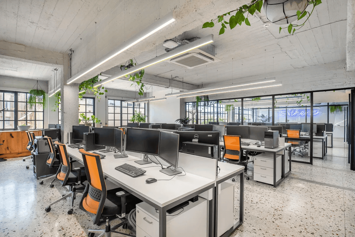 Ferryscanner's open office space with rows of modern white desks, large monitors, and ergonomic orange chairs. The area is well-lit with sleek overhead lighting, hanging plants, and large windows that bring in natural light.
