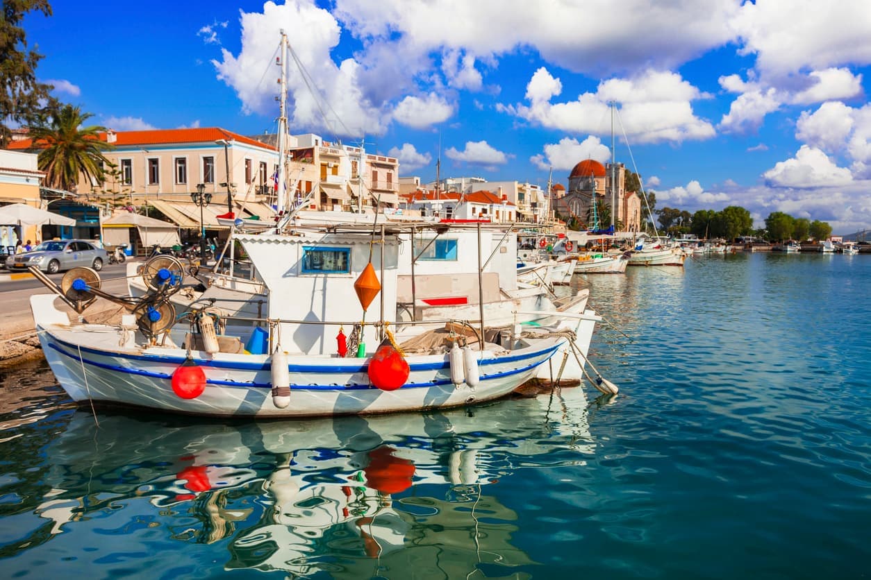 Bateaux de pêche dans le port d'Égine devant des bâtiments traditionnels et une église au dôme rouge.