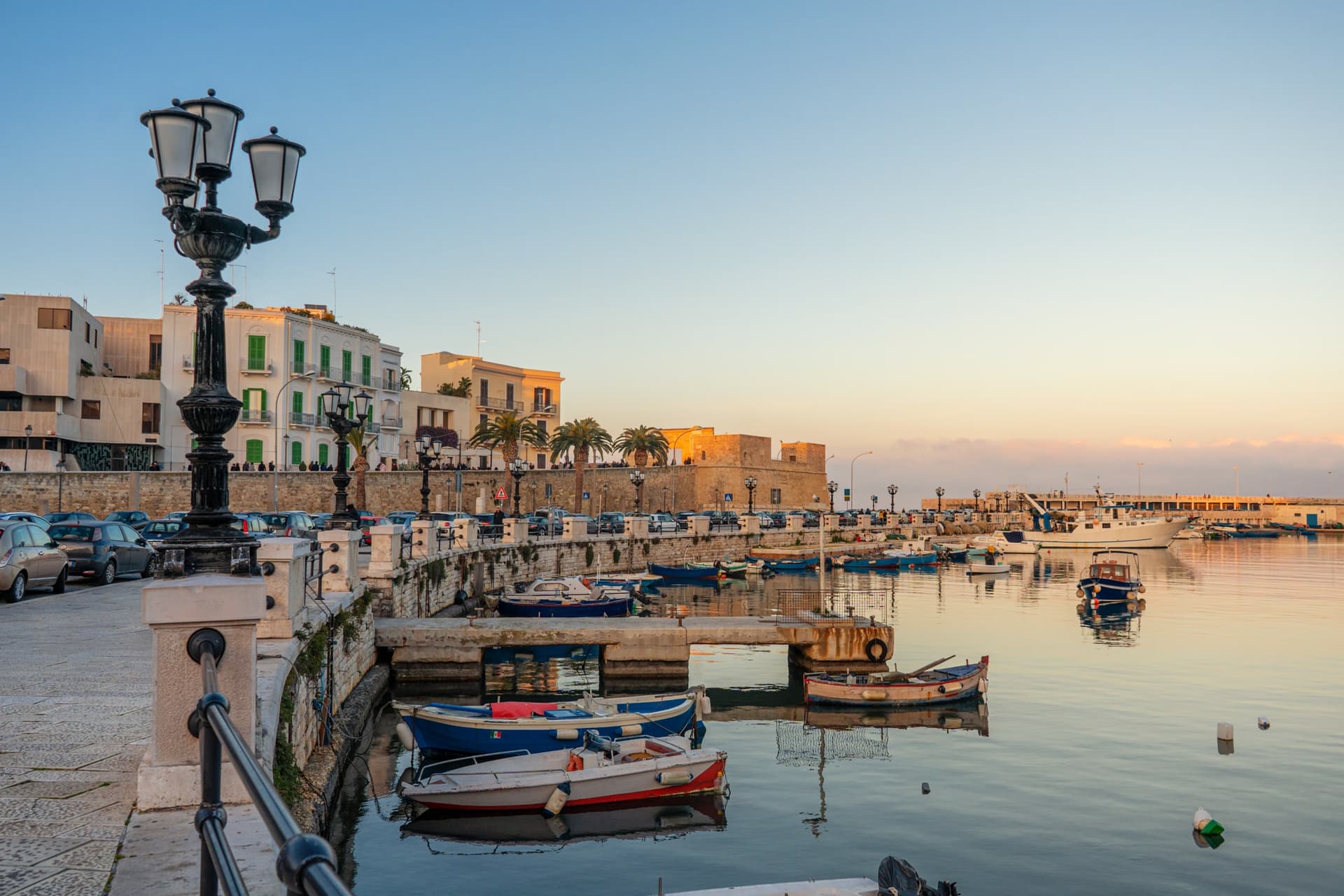 Scenic view of Bari, Italy, with a charming harbor, small boats, historic buildings, and a sunset-lit waterfront.