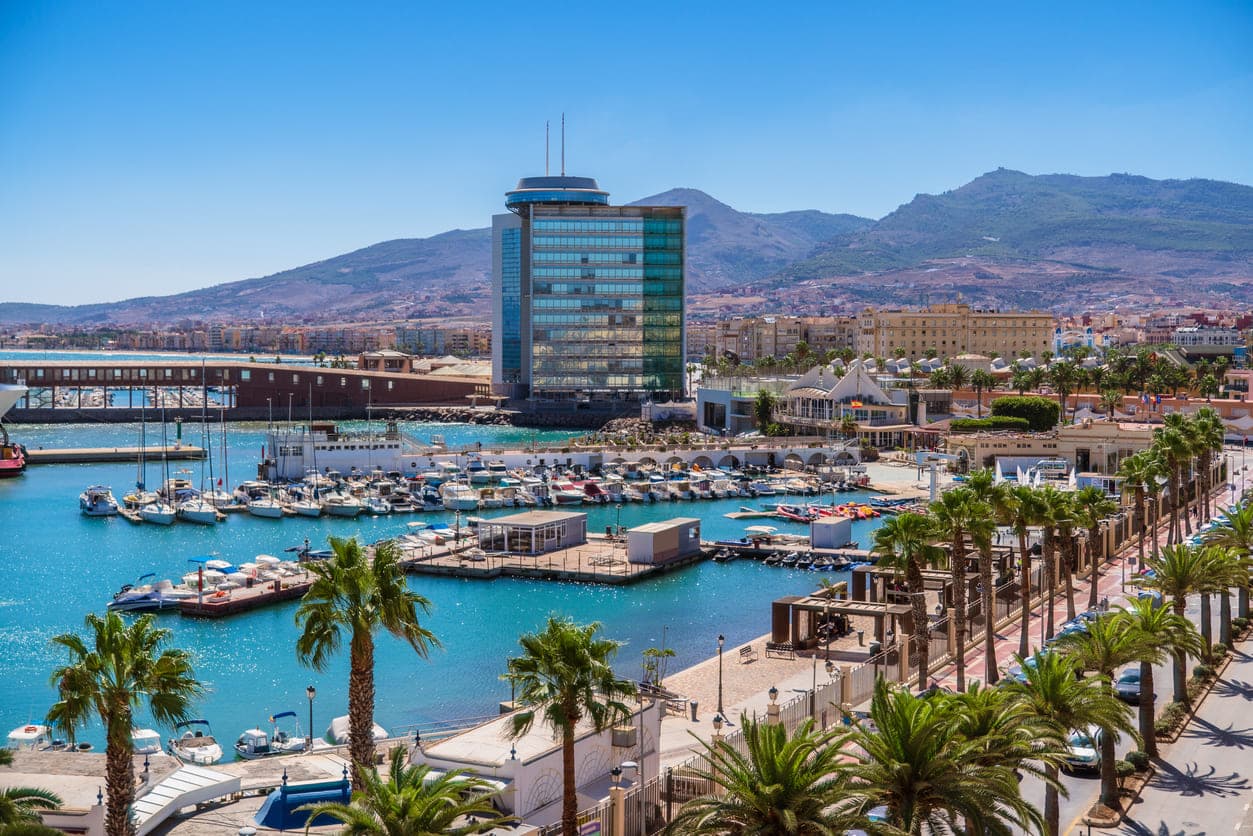 Šetnica Paseo Marítimo uokvirena palmama, brojnim molovima koja vodi do marine marina „Puerto Deportivo Noray“ s velikom staklenom zgradom u španjolskom gradu Melilli u Africi.