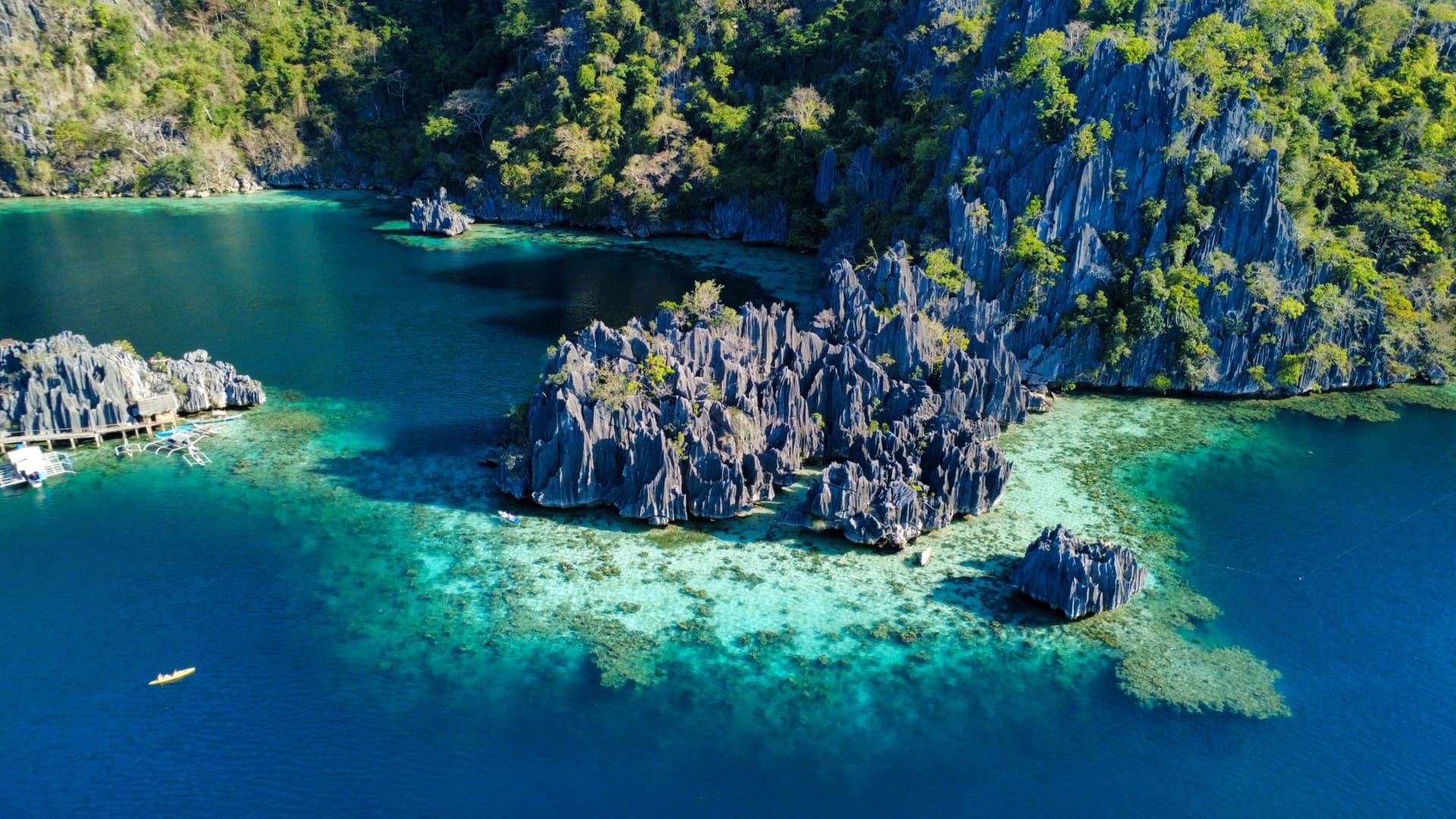 An aerial view reveals a breathtaking tropical lagoon framed by dramatic limestone cliffs and lush greenery. Jagged rock formations rise sharply from crystal-clear turquoise waters, creating striking contrasts between deep blue channels and shallow coral reefs below. Small boats drift quietly across the lagoon, emphasizing the scale and untouched beauty of this remote island paradise. The scene captures a perfect blend of rugged nature and serene coastal charm, inviting exploration and wonder.